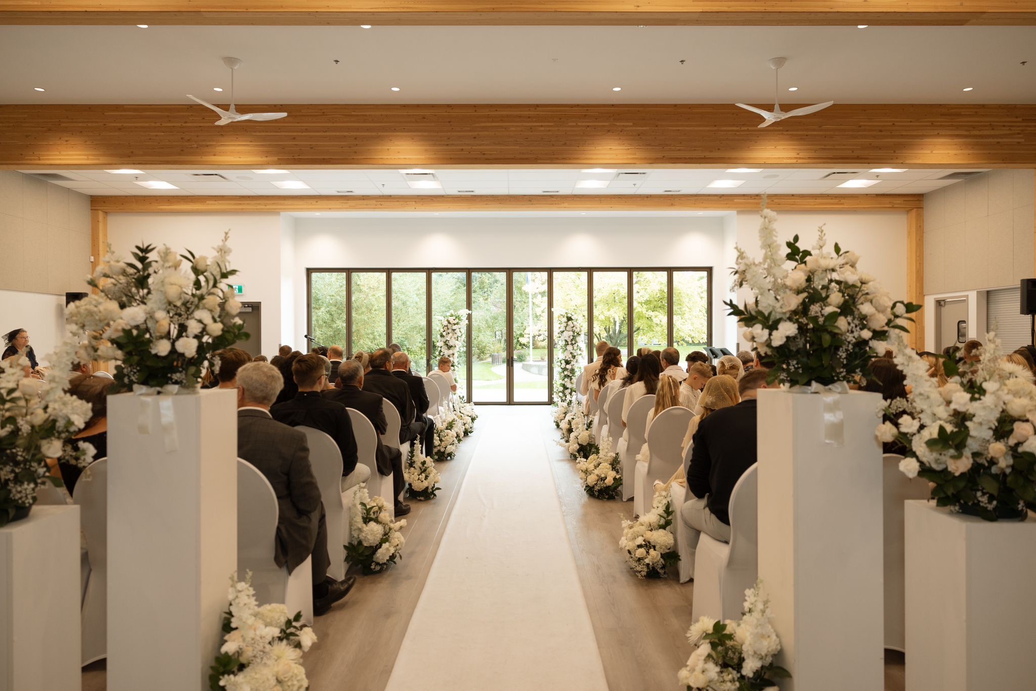 Full ceremony view of a real Okanagan wedding with white faux floral pillars by Kelowna Wedding Arches framing the altar
